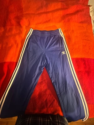 Blå Adidas träningsbyxor med vita ränder - Säljer ett par blå träningsbyxor från Adidas med klassiska vita ränder längs sidorna och Adidas-logga på ena benet. Byxorna har elastisk midja och raka ben, perfekta för en sportig look.