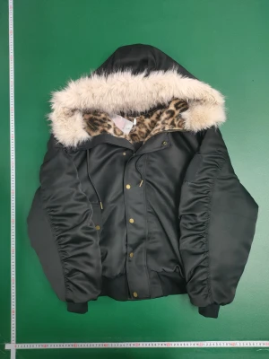 Svart bomberjacka med leopardfoder - Svart bomberjacka med fluffig fuskpäls runt huvan och leopardmönstrat foder. Jackan har militärinspirerad badge på ärmen, guldfärgade knappar och dragkedja. Materialet är nylon och akryl, och modellen är croppad med långa, puffiga ärmar.