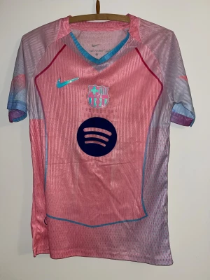 Rosa ”Aurora” FC Barcelona Nike fotbollströja - Säljer en FC Barcelona fotbollströja från Nike i rosa och ljusblå toner. Tröjan har korta ärmar, V-ringad krage med blå kant, och är tillverkad i lätt, ventilerande Dri-Fit material. FCB-logga och Spotify-tryck på bröstet, samt Nike Swoosh i holografisk stil.