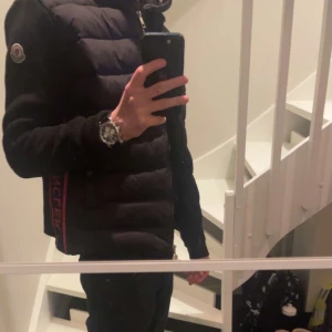 Moncler cardigan - Svart pufferjacka från Moncler med quiltad front och stickade ärmar. Jackan har dragkedja, hög krage och röda samt mörkblå detaljer längs sidorna med Moncler-logga. Materialet är dunfyllning och stickad polyester, perfekt för kyliga dagar.