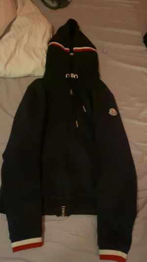 Svart Moncler hoodie med dragkedja  - Säljer en svart hoodie från Moncler med dragkedja och huva. Jackan har vita och röda ränder på huvan och ärmsluten samt Moncler-logga på ärmen. Tillverkad i mjuk bomull och har snörning i huvan. Perfekt för dig som vill ha en clean och stilren look.