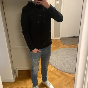 Svart hoodie från Les Deux - Svart hoodie från Les Deux med guld färgad broderad logga på bröstet. Tröjan har huva med snörning, magficka och ribbade muddar vid ärmslut och nederkant. Perfekt för chill dagar och enkel att matcha med jeans eller joggers.