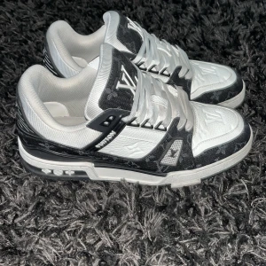 Louis Vuitton svartvita sneakers - Säljer ett par Louis Vuitton sneakers i svart och vitt med tydlig LV-logga på plösen och präglad på tån. Skorna har snörning, platt sula och detaljer i skinn samt textil. Monogrammönster syns på flera partier och sulan har tydlig Vuitton-branding. Riktigt lyxiga och unika sneakers.