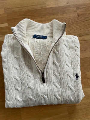 Vit Cable-Knit Quater-Zip Polo Ralph Lauren  - Snygg vit stickad tröja från Polo Ralph Lauren med klassiskt kabelstickat mönster och half zip-dragkedja vid kragen. Tröjan har långärmad passform och den ikoniska broderade loggan på bröstet. Perfekt för dig som gillar stilren och tidlös design. OBS! Det är L men känns mer som M! Annars inga konstigheter. 