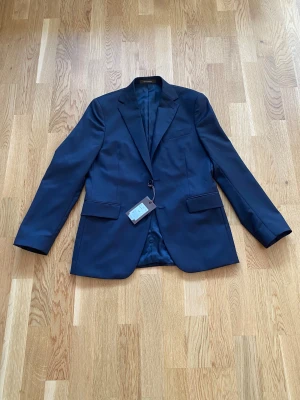 Mörkblå kavaj från Oscar Jacobson - Snygg mörkblå kavaj från Oscar Jacobson med klassisk passform och två framfickor med lock. Kavajen har slät yta, knappar vid ärmslut och är helfodrad med diskret logotyp på insidan. Nypris 4000