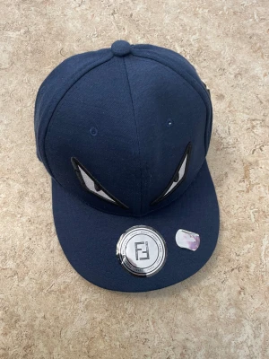 Mörkblå Fendi keps med ögonmotiv - Säljer en mörkblå keps från Fendi med broderade ögon framtill och justerbar snapback baktill. Kepsen har Fendi-logga på baksidan och en klassisk platt skärm. Tillverkad i bomull och perfekt för dig som vill sticka ut med en cool accessoar.
