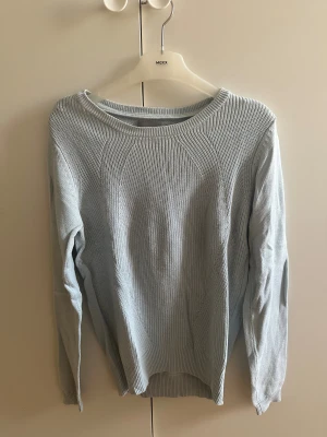 Ljusblå stickad tröja med rund hals - Snygg ljusblå stickad tröja från Vero Moda med rundad hals och ribbade muddar vid ärmslut och nederkant. Tröjan har ett diskret mönster framtill och är långärmad, perfekt för dig som gillar enkel och stilren design