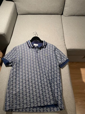 Blå mönstrad pikétröja Lacoste M - Snygg blå pikétröja från Lacoste med geometriskt mönster i vitt. Klassisk krage med vita ränder och knappar med Lacoste-logga. Tröjan har korta ärmar och normal passform. Perfekt för dig som gillar stilrena detaljer och ikonisk design.