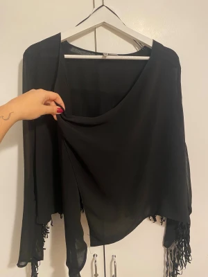Svart transparent jacka  - Säljer en svart jacka i transparent, lätt material med långa ärmar och snygga fransdetaljer längs ryggen och ärmarna. Blusen har en lös passform och asymmetrisk nederkant som ger en cool och unik look.