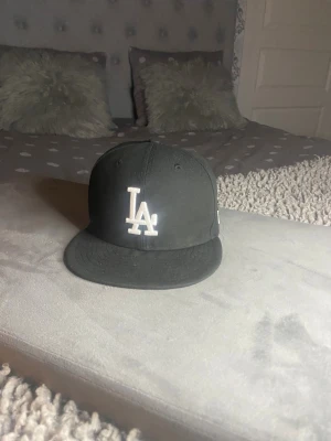Svart LA-keps från New Era - Säljer en svart keps från New Era med LA Dodgers-logga broderad i vitt framtill och MLB-logga bak. Klassisk flat brim och strukturerad passform. Kepsen är tillverkad i bomull och polyester, perfekt för dig som gillar sportig streetstyle.