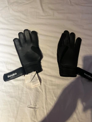 Sondico svarta målvaktshandskar - Två par svarta målvaktshandskar från Sondico. Ena paret är helsvart med breda kardborreband och Sondico-logga, det andra paret har svart bas med gula detaljer och Sondico S på ovansidan. Båda paren har förstärkta fingrar och greppvänlig insida. Prövat dom en gång men dom var för små för mig 