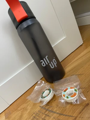 Air Up vattenflaska med smakpods - Snygg Air Up vattenflaska i svart med röd detalj, inklusive två oöppnade smakpods (apelsin och ananas). Flaskan och pods är i mycket gott skick, inga synliga repor eller skador. Perfekt för dig som vill dricka mer vatten med smakupplevelse utan tillsatt socker. Flaskan och podden kommer med. Använts men är som ny 