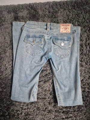 True Religion ljusblå bootcut jeans - Säljer ett par ljusblå bootcut jeans från True Religion med snygga slitningar framtill och ikoniska fickor med lock och silverknappar bak. Jeansen har låg midja och klassisk femficksdesign. Materialet är mjukt jeans och passformen är loose.