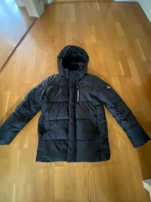  Calvin Klein jacka  - Svart pufferjacka från Calvin Klein Jeans med huva och diskret logga på ärmen. Jackan har dragkedja framtill och två fickor med dragkedja. Perfekt för kalla dagar och har en stilren, enkel design som funkar till det mesta.