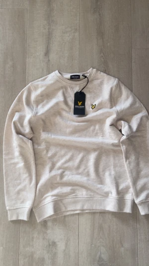 Beige sweatshirt från Lyle & Scott - Snygg beige sweatshirt från Lyle & Scott med klassisk rund hals och gul broderad logga på bröstet. Tröjan har ribbade muddar vid ärmslut och nederkant, samt en diskret V-detalj vid halsen. Perfekt för en chill och stilren look.