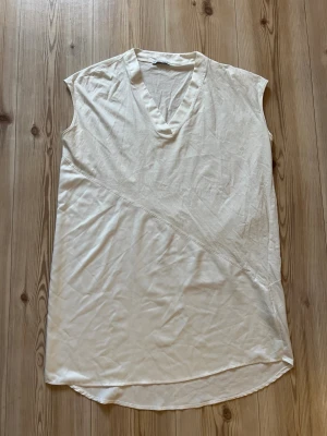 Vit tunika från Zara - Vit tunika från Zara med v-ringning och asymmetrisk söm framtill. Klänningen har en rak, loose passform och är tillverkad i ett mjukt, lätt material som känns svalt mot huden. Perfekt för varma dagar och enkel att styla med accessoarer.