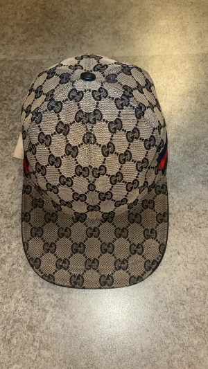 Gucci keps - Säljer en beige keps från Gucci med klassiskt GG-monogram i svart över hela kepsen. Kepsen har en böjd skärm och justerbart spänne bak. På sidan finns en ikonisk röd och marinblå rand. Materialet är bomullscanvas med insida i mörkblått tyg.