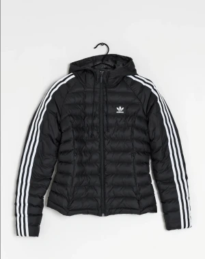 Svart pufferjacka från Adidas - Svart pufferjacka från Adidas med klassiska vita ränder längs ärmarna och Adidas-logga på bröstet. Jackan har huva, dragkedja framtill och är quiltad för extra värme. Perfekt för kyliga dagar och har en sportig vibe.