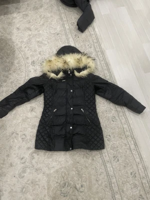 Svart Rockandblue dunjacka med äkta päls - Svart lång dunjacka från Rockandblue med quiltade partier och snygg äkta päls på huvan. Jackan har dragkedjor, knappar och flera fickor, samt ett Rockandblue-märke på axel. Perfekt för kalla dagar och riktigt stilren look.