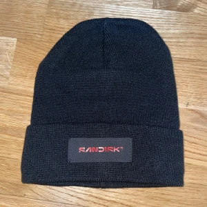 Svart stickad mössa från SanDisk - Svart, klassisk stickad mössa med uppvikt kant och SanDisk-logga i rött framtill. Enkel och stilren design som passar perfekt till kalla dagar. Tillverkad i mjukt material för skön känsla.