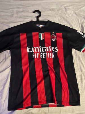 AC Milan Ibrahimović matchtröja - Snygg AC Milan fotbollströja med röda och svarta ränder, officiella klubbmärket och sponsortryck. På ryggen står Ibrahimović och nummer 11. Korta ärmar med italienska flaggans färger på ärmslut. Tillverkad i lätt och ventilerande polyester.