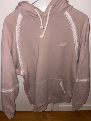 Beige hoodie från Kith med broderi - Säljer en beige hoodie från Kith med vit broderad logga på bröstet och dekorativa vita detaljer längs sömmarna. Tröjan har huva med snörning, stor ficka framtill och är tillverkad i mjuk bomull. Perfekt för chill dagar och snygg att matcha med jeans.