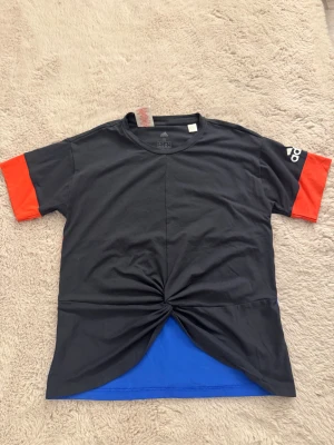 Adidas t-shirt  - Cool t-shirt från Adidas i svart, rött och blått och adidas logga på ena ärmen. Tillverkad i Climalite-material som andas och minskar risken för svett. Snygg twistad detalj framtill och breda färgblock på ryggen. Perfekt för dig som gillar sportig och unik stil!! (Aldrig använd- nyskick) 