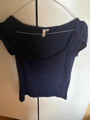 Mörkblå topp med spetskant från Nelly - Säljer en mörkblå topp från Nelly i storlek M. Toppen har korta ärmar, en bred v-ringning och en söt spetskant längs halsringningen. Perfekt att styla med jeans eller kjol för en enkel men snygg look.