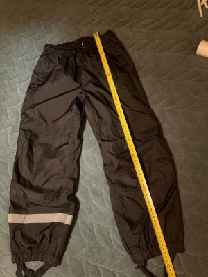 Svarta skalbyxor  med reflexdetalj - Svarta skalbyxor från Bik Bok med elastisk midja och justerbara benslut. Utrustade med reflexdetalj på ena benet och praktiska fothällor. Yttermaterialet är syntet och insidan har meshfoder för extra komfort. Perfekta för kalla och blöta dagar.