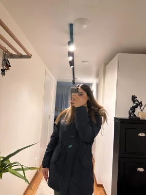 Mörkblå parkas från Lyle & Scott - Parkas i mörkblått från Lyle & Scott med fuskpäls på huvan, snörning i midjan och dragkedja framtill. Jackan har stora fickor och klassisk gul logga på bröstet. Perfekt för kalla dagar och snygg till streetwear.