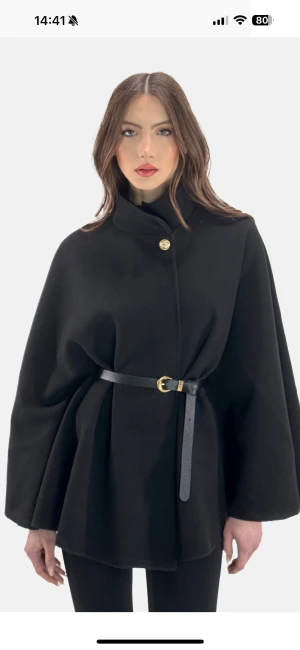 Svart poncha med bälte och guldknapp  - Svart kappa med vid, svepande passform och bälte i midjan med gulddetalj. Kappan har en hög krage och en dekorativ guldknapp upptill. Tidlös och elegant modell med vida ärmar, perfekt för lager-på-lager. Materialet ser ut att vara ullblandning.