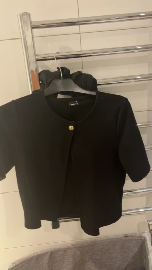 Svart kofta  från Gina Tricot med guldfärgad knapp - Säljer en stilren svart kofta från Gina Tricot med korta ärmar och rund halsringning. koftan  har en snygg guldfärgad knapp framtill och är gjord i ett mjukt material som känns skönt mot huden. Perfekt att styla över en t-shirt eller linne.