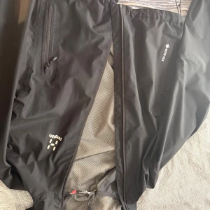 Svart vindjacka från Haglöfs Gore-Tex - Svart vindjacka från Haglöfs med Gore-Tex-material, perfekt för blåsiga dagar. Jackan har dragkedja framtill, meshfoder på insidan och diskreta loggor på bröstet. Snygg och funktionell design med fickor med dragkedja.