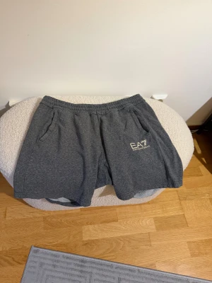 Grå EA7 shorts med logga - Mörkgrå shorts från EA7 Emporio Armani med elastisk midja och sidofickor. Mjuk bomullskänsla och vit EA7-logga på ena benet. Perfekta för chill eller träning, klassisk och enkel design.