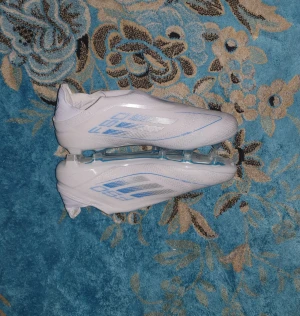 Adidas F50 vita fotbollsskor - Säljer ett par Adidas F50 fotbollsskor i vitt med blå och silver detaljer. Skorna har en strömlinjeformad design utan snörning, med platt sula och rund tå. Yttermaterialet är syntet och sulan har blå inslag för extra stil. Perfekt för dig som vill sticka ut på planen. De passar storlek 44-45 