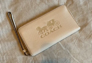 Beige plånbok från Coach med handledsrem - Snygg beige plånbok från Coach med präglad logga och häst-vagn motiv på framsidan. Plånboken är rektangulär, har en praktisk handledsrem och stängs med dragkedja i guld. Tillverkad i strukturerat skinn som ger en lyxig känsla.