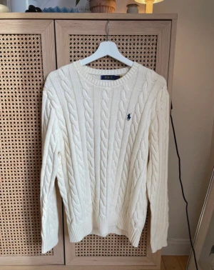 Polo Ralph Lauren - cable Shirt  - Snygg vit kabelstickad tröja från Polo Ralph Lauren med klassisk blå broderad logga på bröstet. Rund hals, ribbade muddar och långärmad. Perfekt för dig som gillar clean och tidlös stil. Materialet är mjuk bomull som känns skönt mot huden.