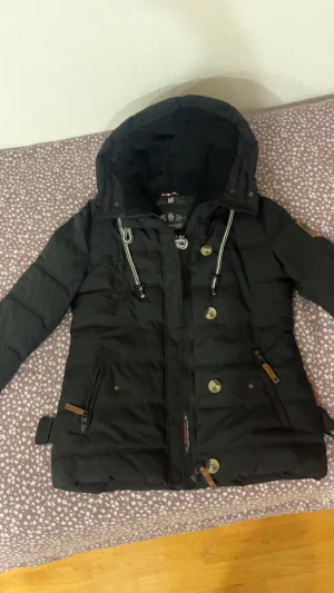 Svart pufferjacka med huva från Navigare - Svart pufferjacka från Navigare med huva, snörning och dragkedja samt stora knappar framtill. Jackan har långa ärmar, två fickor med dragkedja och detaljer i läderimitation. Perfekt för kalla dagar och riktigt snygg passform.