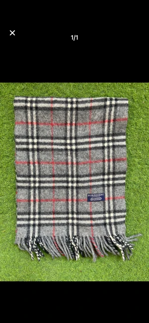 Rutig grå halsduk från Burberry - Klassisk halsduk från Burberry i 100% kashmir. Grå bas med rutigt mönster i svart, vitt och röda linjer. Mjukt material och fransar i ändarna. Perfekt accessoar för att lyfta din outfit med en tidlös brittisk vibe.