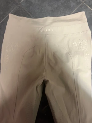 Beige ridbyxor från Equiline - Snygga beige ridbyxor från Equiline med hög midja och tight passform. Byxorna har diskret broderad logga och dekorativa detaljer på sidorna. Materialet är stretchigt och bekvämt, perfekt för ridning. Stilren design med broderade mönster för extra touch.