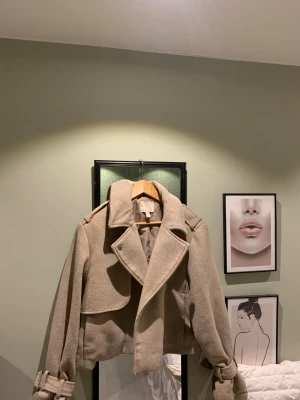 Beige croppad skepparkavaj från H&M - Snygg croppad skepparkavaj i beige från H&M. Jackan har bred krage, markerade axlar och bälte vid ärmsluten för extra edge. Tillverkad i ullmix med slät insida. Perfekt för dig som vill ha en stilren och trendig look.
