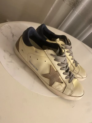 Golden Goose Superstar sneakers - Storlek 42, Golden Goose Superstar sneakers i beige skinn med grå mockastjärna på sidan, grå snörning och mörkblå häl. Klassisk låg modell med platt sula och cool vintagekänsla. Perfekta för dig som gillar streetstyle och unika detaljer. Dem har blivit lite gula 