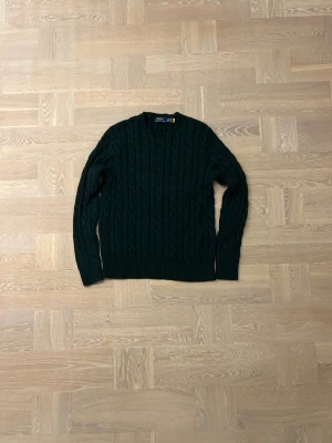 Mörkgrön stickad tröja från Polo Ralph Lauren - Snygg mörkgrön stickad tröja från Polo Ralph Lauren med klassiskt flätmönster. Tröjan har rund hals, ribbade muddar och långärmad passform. Perfekt för dig som gillar stilrena och tidlösa plagg med lite extra struktur. Pris kan diskuteras vid snabb affär