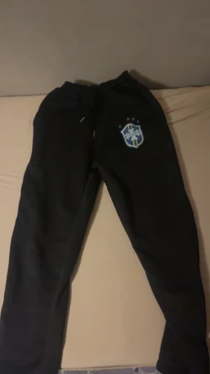 Svarta mjukisbyxor med Saint-logga - Svarta mjukisbyxor med broderad Saint-logga och stjärnor på vänster ben. Byxorna har snörning i midjan och är gjorda i mjukt material som passar perfekt för chill eller sport. Klassisk passform med elastisk midja för extra komfort.