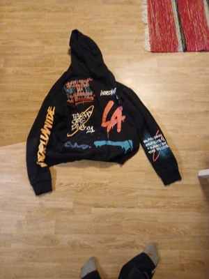 Svart hoodie Black Squad med graffiti - Svart hoodie från Black Squad i relaxed fit, storlek L. Tröjan har färgglada graffiti-prints och text på både fram- och baksida samt ärmar. Huva och muddar vid ärmslut och nederkant. Perfekt för dig som gillar streetstyle och unika detaljer.