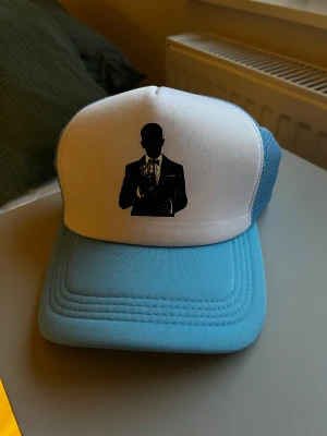 Vit och ljusblå truckerkeps med tryck Luxestrap - Snygg truckerkeps i vit och ljusblå färg med mesh på sidorna och baksidan. Framtill finns ett svart tryck av en siluett i kostym som håller pengar. Kepsen har justerbar snapback och är tillverkad i polyester och syntetmaterial.