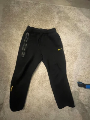 Svarta Nike mjukisbyxor med gula detaljer - Svarta mjukisbyxor från Nike med gul swoosh-logga och gula tryckdetaljer på benen. Byxorna har snörning i midjan och är tillverkade i mjukt bomullsmaterial. Perfekta för chill eller träning, med en loose fit och sportig vibe.