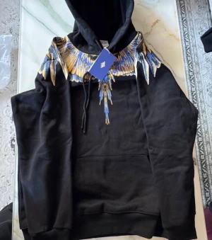 Marcelo Burlon svart hoodie med vingar - Svart hoodie från Marcelo Burlon County of Milan med ikoniskt vingtryck i blått och gult över axlar och rygg. Tröjan har huva med snörning och stor magficka framtill. Perfekt för dig som vill sticka ut med en fet design och streetwear-vibe.
