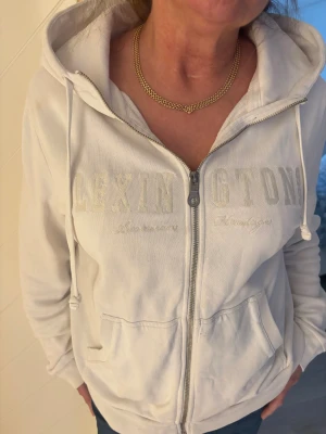 Vit hoodie från Lexington med dragkedja - Säljer en vit hoodie från Lexington med broderad logga på bröstet. Tröjan har huva med snörning, praktiska fickor och dragkedja framtill. Tillverkad i mjuk bomull och har långa ärmar. Liten fläck på armen, går säkert bort med galltvål. Se bild 2. Nypris 1600kr. Storlek L men sitter bra på mig som är en M