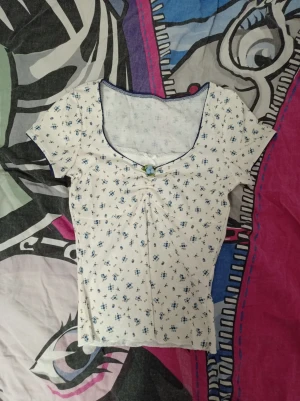 Vit blommig topp med rosett och spets - Supersöt vit topp med små blå blommor, korta ärmar och v-ringning. Dekorerad med en liten blå rosett och spetskant vid halsen. Toppen har rynkdetalj framti .obs denna tröja sitter ganska tight på mig som är xs eller xss